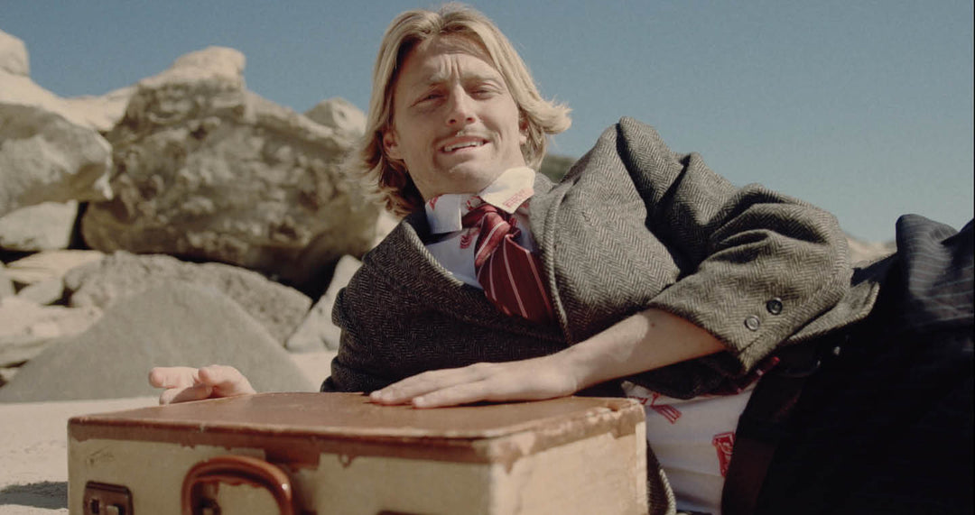 JOSIAH AMICO laying on the beach in a suit with a wooden box in front of him