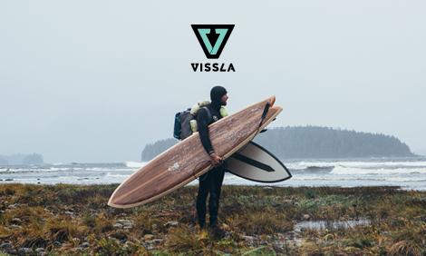 man in hooded wetsuit standing on grass with a backpack and two surfboards, looking out at the waves